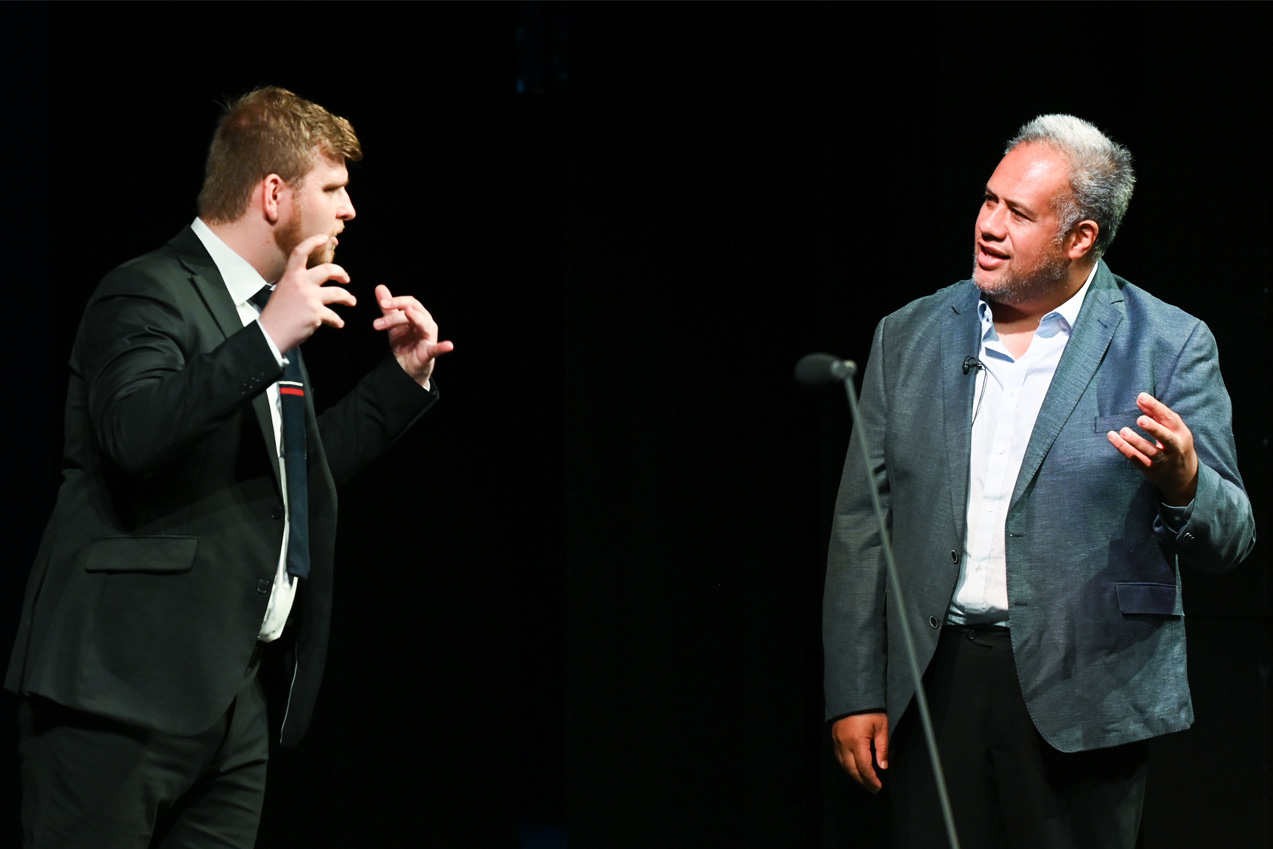 A male student, wearing a black tie and suit, talking on stage, with a man, wearing a white shirt and dark blazer, in a masterclass lesson